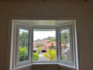 View of a residential street through uPVC double glazed windows in Bondi, with greenery and red-roofed houses visible in the background.