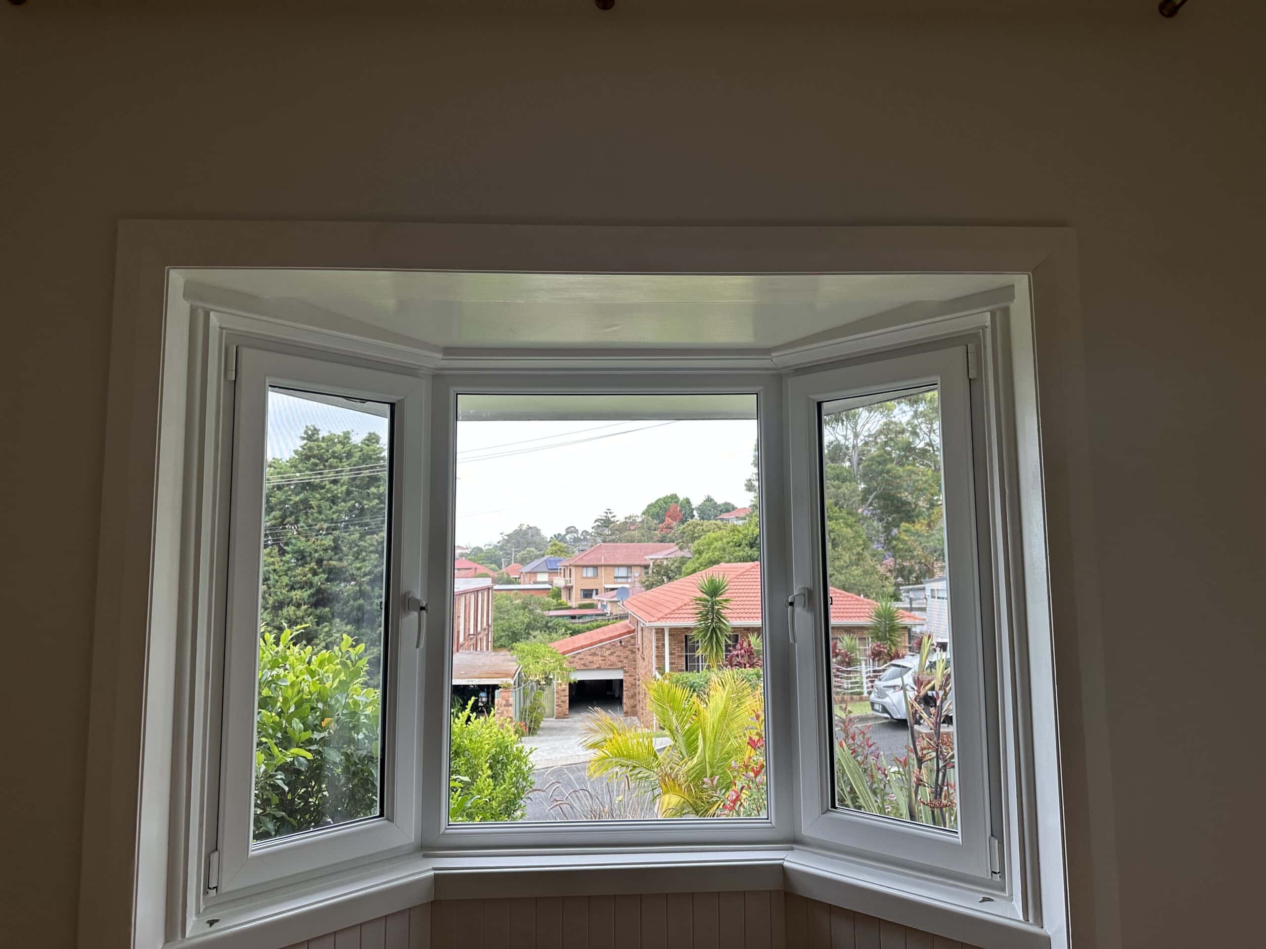View of a residential street through uPVC double glazed windows in Bondi, with greenery and red-roofed houses visible in the background.