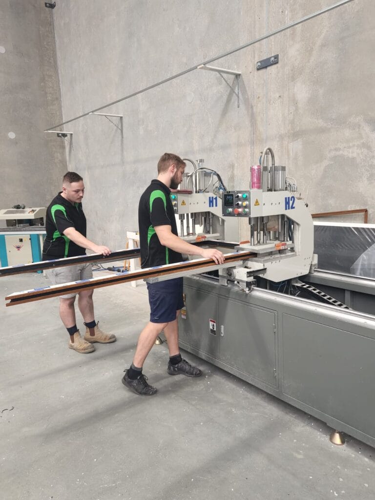 In a partnership with Solace Creations, two skilled workers expertly operate a large industrial cutting machine in the workshop, diligently handling long wooden panels.