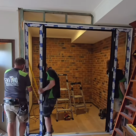 Two workers are installing a large UPVC door frame in a room with brick walls, perhaps hinting at a future wine cellar. Equipment such as ladders and tools are visible in the background, ready to assist with every fine detail.