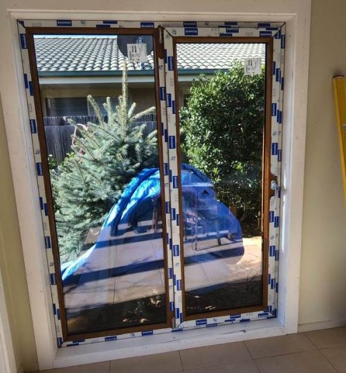 UPVC French & Single Hinge Doors 2 Newly installed glass sliding doors with blue protective tape, reflecting a partially covered outdoor area with greenery and a conifer tree.
