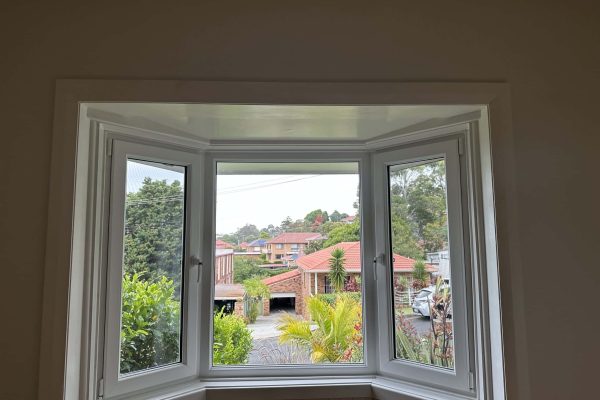 View of a residential street through uPVC double glazed windows in Bondi, with greenery and red-roofed houses visible in the background.