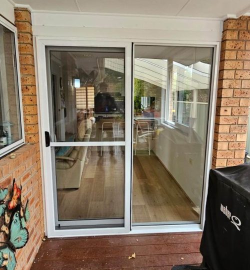 UPVC Sliding and Stacking Doors 1 A sliding glass door on a porch opens to a well-lit interior room with wooden flooring. A grill is present on the right, and decorative items are attached to the brick wall on the left.